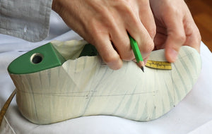 Person measuring a wooden shoe insole with a green tool on a white surface. VALEVRO footwear design course.