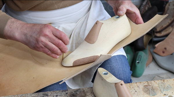 A close-up of a leather shoe molding process, showing a craftsman's hands shaping a light tan leather insole around a wooden foot mold held over a large piece of tan leather.
