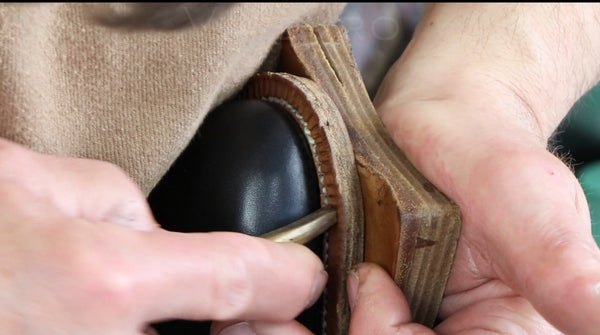 A close-up of a person's hands holding a black leather shoe or boot while using a wooden and metal tool to shape or polish the edge of the leather.