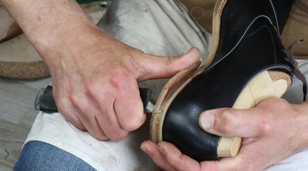 A close-up of a person's hands holding a black leather shoe with a light-colored wooden foot block, using a tool to trim or shape the edge of the shoe.