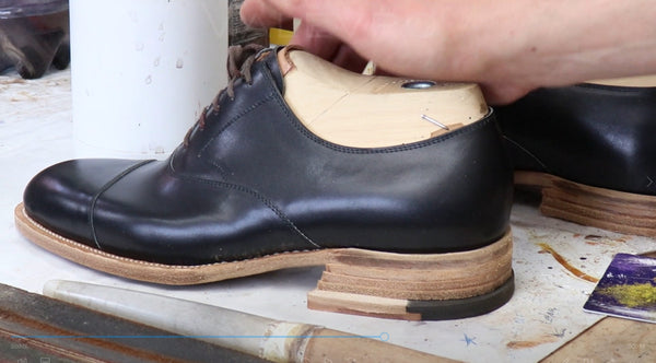 A close-up of a black leather dress shoe with a wooden shoe tree inserted, held by a hand in a workshop setting.