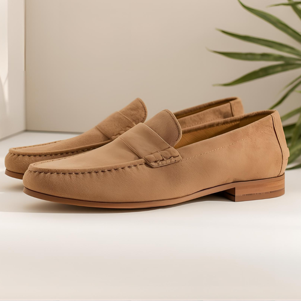 A light brown suede leather loafer, clean and minimal background in pale tones, with a soft light green plant slightly visible in the corner.