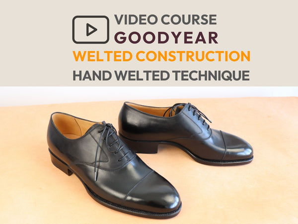 A pair of black leather cap-toe oxford shoes with a polished finish, displayed on a light beige surface below a text banner that reads 'VIDEO COURSE GOODYEAR WELTED CONSTRUCTION HAND WELTED TECHNIQUE'.